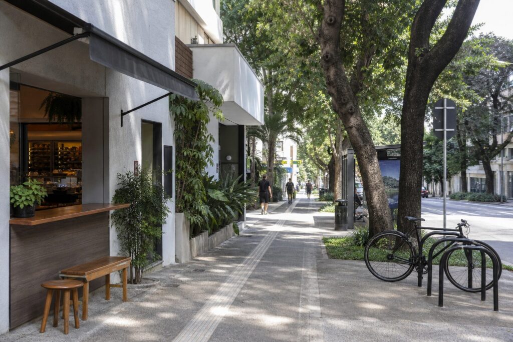Calçada arborizada de rua residencial em São Paulo, com comércio de bairro e bicicletário